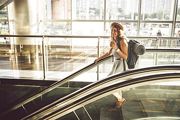 Portrait Smiling Woman Wearing Hat In Airport At Escalator. people travelling with hand luggage. Theme tourism and transport. Caucasian girl with coffee in airport terminal on an escalator