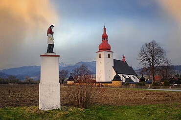 Church of St. Ladislaus in Liptovske Matiasovce at sunset in Liptov with a statue of a saint by the road