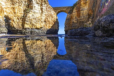 Yellow rocks and cliffs on a sandy beach. A bay by the sea, over which the old stone bridge of the Pinhão fortress connects two cliffs. Praia da Caldeira, Lagos. Algarve, Portugal, Europe