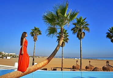 Beautiful woman standing on bent palm tree trunk at infinity pool long red dress