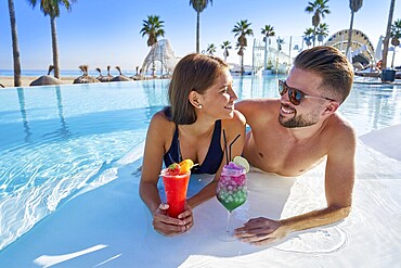 Young tourists couple on infinity pool drinking cocktails at resort on the beach