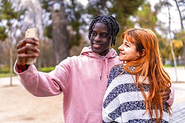 Young multiracial couple smiling together while capturing a joyful selfie with a smartphone in a vibrant park setting