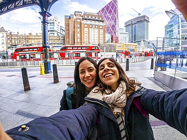 Two happy tourists taking a selfie in the city of london, with red buses and modern buildings in the background
