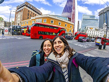 Two happy female tourists taking a selfie with red double decker buses and modern skyscrapers in the background, enjoying their vacation in london
