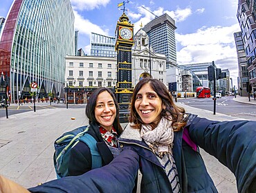 Two happy tourists taking a selfie in london near a historical clock tower, with modern buildings and red bus in the background