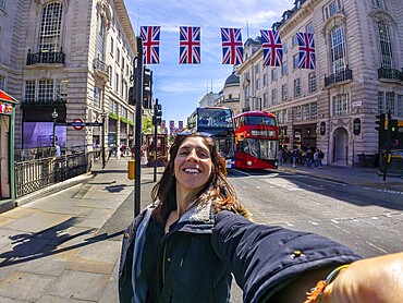 Young woman taking a selfie in london with union jack flags and a red double decker bus in the background, enjoying a sunny day