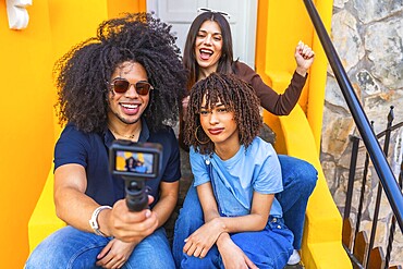 Three happy multi ethnic vloggers recording a video blog outdoors, sitting on colorful yellow stairs