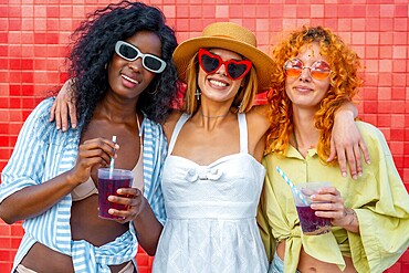 Three multi ethnic young women hugging each other and drinking juice with straw, standing against a red tiled wall