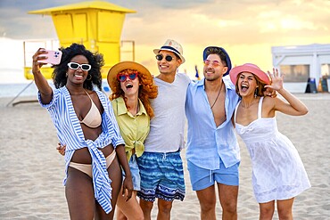 Group of multi ethnic friends taking a selfie on the beach in miami, florida, with lifeguard tower and sunset in background