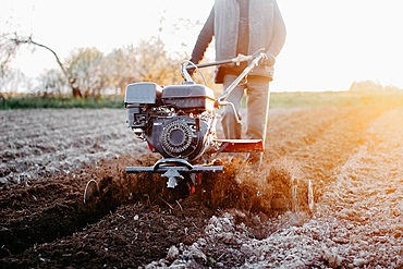 Person using a tiller on a farm field at sunrise, emphasizing farming and manual labor