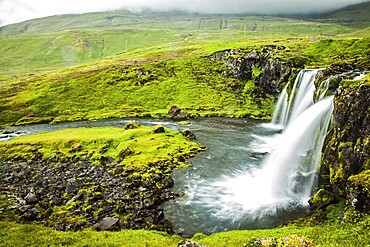 Overcast cold summer day in Iceland. Concept of extreme and exotic tourism. At the foot of the mountain Kirkjoufell powerful waterfalls Kirkjoufellfoss