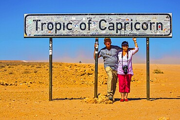 Happy elderly couple of tourists. South Tropic, or Tropic of Capricorn, - the famous road sign in Namibia. The concept of extreme and active tourism