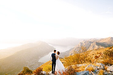 The bride and groom stand on the top of Mount Lovcen overlooking the Bay of Kotor, smile and hug tenderly .