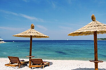 Two wooden loungers on the white sand, on a paradise beach near the town of Primosten in Croatia. Straw beach umbrella. Blue sky and azure Adriatic Sea. The concept of beach holidays and travel