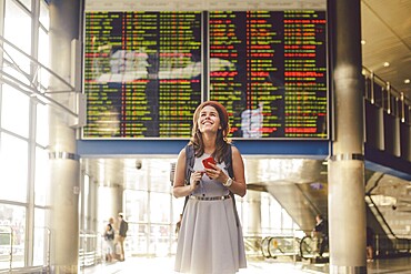 Theme travel and transport. Beautiful young caucasian woman in dress and backpack standing inside train station or terminal looking at a schedule holding a red phone, uses communication technology