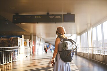 Theme travel public transport. young woman standing with back in dress and hat behind backpack and camping equipment for sleeping, insulating mat looks schedule on scoreboard airport station sunny day
