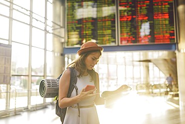 Theme travel and transportation. Beautiful young caucasian woman in dress and backpack standing inside train station terminal looking at electronic scoreboard holding phone, map paper hand navigation