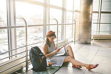 Theme of tourism and travel of young student. Beautiful young caucasian girl in dress and hat sits on floor tourist rug inside terminal airport terminal. Waiting room delayed flight, delay departure