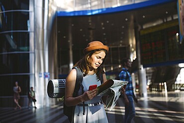Theme travel and transportation. Beautiful young caucasian woman in dress and backpack standing inside train station terminal looking at electronic scoreboard holding phone, map paper hand navigation