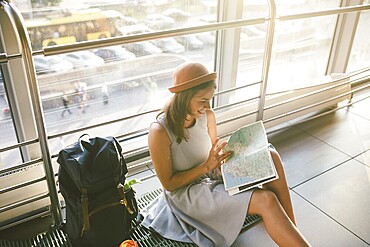Waiting, delayed transport in the terminal of the airport or train station. Young caucasian woman in dress and hat sits on tourist rug with backpack near window to room and looks into tourist map