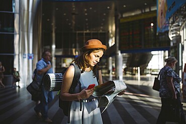 Theme travel and transportation. Beautiful young caucasian woman in dress and backpack standing inside train station terminal looking at electronic scoreboard holding phone, map paper hand navigation