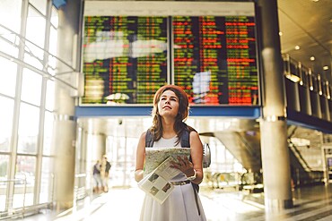 Theme travel and transportation. Beautiful young caucasian woman in dress and backpack standing inside train station terminal looking at electronic scoreboard holding phone, map paper hand navigation