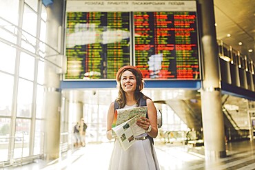 Theme travel and transportation. Beautiful young caucasian woman in dress and backpack standing inside train station terminal looking at electronic scoreboard holding phone, map paper hand navigation
