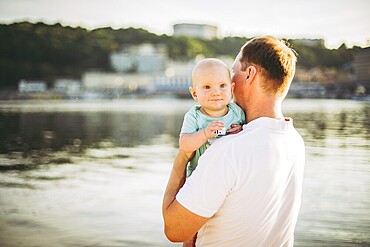 Subject Parenting, summer holidays, father and little son. Young Caucasian dad holds on arms, hugs child overlooking town Kiev and the river on banks of reservoir, overlooking lake at sunset