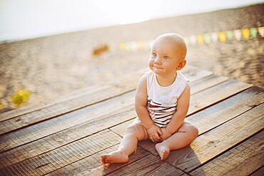 Child boy one year blond sits on a wooden dock, a pier in striped clothes, a compound near the pond on a sandy beach against a background of a river in the summer at sunset of the day