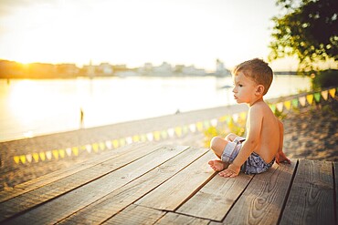 The theme is child and summer beach holiday. A small Caucasian boy sits sideways on a wooden pier and admires the view of the sandy beach and the pond, the river. With bare legs in blue denim shorts