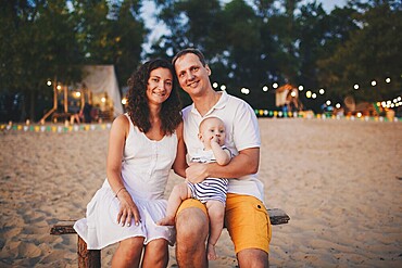 Theme of family summer holidays on the beach. Caucasian man and woman dad and mom hug a small baby son sitting on his hands. Young family relaxing in a beach cafe at sunset