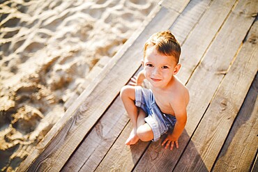 The theme is a child and summer beach holiday. A small Caucasian boy sits sideways on a wooden pier and looks at the camera on a sandy beach and a pond, a river. With bare legs in blue denim shorts