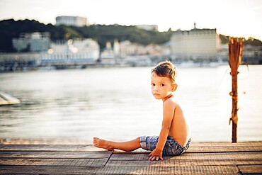 The theme is a child and summer beach holiday. A small Caucasian boy sits sideways on a wooden pier and looks at the camera on a sandy beach and a pond, a river. With bare legs in blue denim shorts