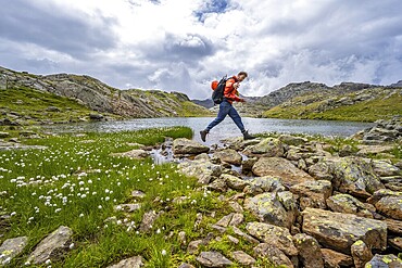 Mountaineer jumping from stone to stone, at a mountain lake in high mountain landscape at the Obere Senner Egete, Seven Lakes Hike, Stubai Alps, near Ridnaun, South Tyrol, Italy