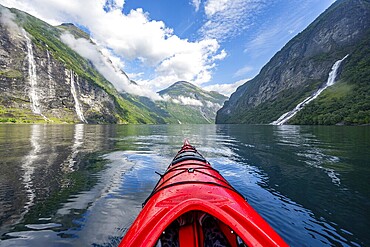 Kayak trip on the Geirangerfjord with waterfall The Seven Sisters and Freier Waterfall, tip of a red kayak in the fjord, atmospheric fjord landscape with reflection, first person view, near Geiranger, Møre og Romsdal, Norway
