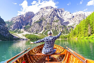 A man sits in a wooden boat and spreads out his arms as he heads towards an impressive mountain, Pragser Wildsee, South Tyrol, Dolomites, Italy