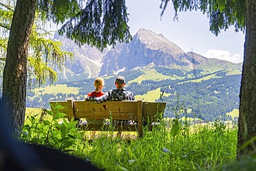 Couple on a bench in front of a wide mountain and forest landscape, Alpe di Siusi, Dolomites, South Tyrol, Italy
