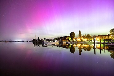 Colourful northern lights reflected in a quiet night lake next to illuminated buildings, harbour, Allensbach, Lake Constance, Baden-Württemberg, Germany