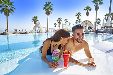 Young tourist couple on infinity pool drinking cocktails at resort on the beach