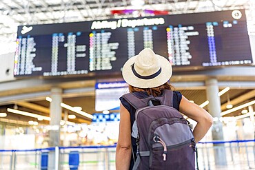 A woman wearing a straw hat and a backpack is standing in front of a large airport display. She is waiting for her flight, possibly at an airport
