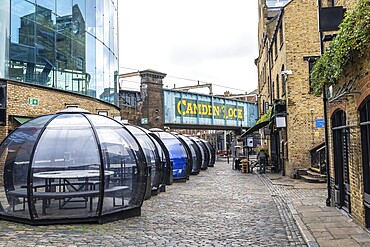 Transparent dining pods line a cobblestone street beneath the Camden lock bridge in London, offering a unique and sheltered dining experience