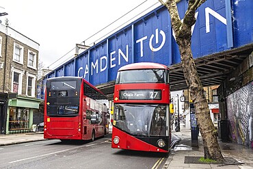 Red double decker buses driving on a London street, passing under a bridge with the Camden town sign