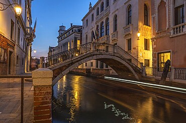 Small pedestrian bridge arching over canal in venice, Italy at twilight with light trails reflecting in water from passing boat