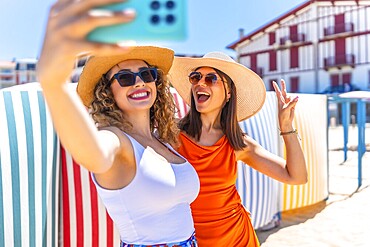 Two cheerful women wearing sun hats and sunglasses, capturing a fun selfie on a sunny beach, radiating joy and friendship