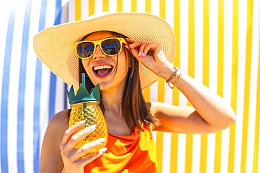 Young woman with sunglasses and a straw hat, enjoying a tropical pineapple juice on a colourful striped background