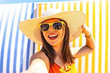 Young woman with sunhat and sunglasses taking a selfie on a striped background at the beach during summer vacation