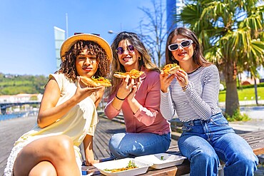 Three women enjoying pizza slices while sitting on a bench in a sunny city park, sharing laughter and savouring a delightful summer day
