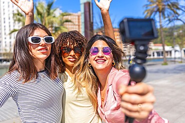Three cheerful women taking a selfie with action camera, waving and smiling, enjoying a summer day together in the city