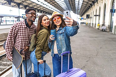 Three happy young tourists taking a selfie with a smartphone while waiting for their train, ready for their next adventure