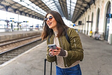 Young woman tourist using her smartphone while waiting for a train at the railway station, smiling and enjoying her travel adventure